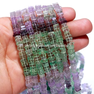 Könnte beinhalten: Eine Strähne aus grünen, violetten und rosafarbenen Fluoritperlen. Die Perlen sind facettiert und haben eine quadratische Form. Die Perlen sind auf einer durchsichtigen Gummikordel aufgefädelt.