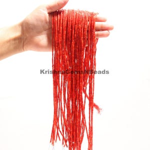 Op de afbeelding: Een hand houdt strengen kleine, rechthoekige, helderrode kralen vast. De kralen zijn geregen en vormen verticale lijnen. De tekst "KrishnaGemsNBeads" is zichtbaar in de afbeelding.