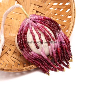 May include: Strands of ruby gemstone beads in varying shades of red and pink, with gold-colored accents. The beads are arranged in a cascading pattern, with a white string and gold thread at the top. The beads are resting on a white stone.