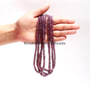 May include: A strand of purple gemstone beads, each bead is a rectangular tube shape. The beads are strung on a clear thread and are held in a hand.