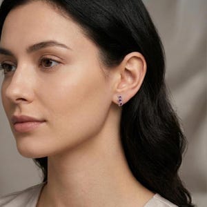 Puede incluir: Primer plano de una oreja con tres pequeños pendientes de botón de piedras preciosas moradas. Los pendientes están engarzados en metal plateado o blanco. La persona tiene el pelo oscuro y un tono de piel neutro.