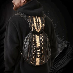 Könnte beinhalten: Ein schwarzer Rucksack mit strukturierter Oberfläche, geflochtenen Lederdetails, goldfarbenen Totenkopf-Akzenten aus Metall, runden Ösen und vertikalen Reißverschlüssen. Das Design umfasst ein Mittelteil, das mit zahlreichen kleinen goldfarbenen münzähnlichen Verzierungen und hängenden Metallflügel-Anhängern versehen ist.