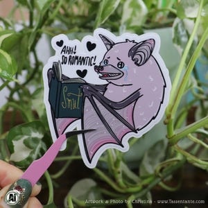 Könnte beinhalten: Ein Aufkleber mit einer Comic-Fledermaus, die ein Buch mit dem Titel "Smut" liest. Die Fledermaus ist helllila mit dunkelgrauen Flügeln und weint. Der Text "AHH! SO ROMANTIC!" und schwarze Herzen sind darüber.