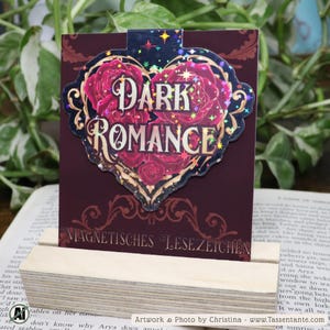 Könnte beinhalten: Burgunderfarbener Lesezeichen mit herzförmigem Design und dem Schriftzug "DARK ROMANCE" in goldfarbenen Buchstaben. Das Herz ist mit roten Rosen gefüllt und mit goldenem Filigran verziert. Der Text "MAGNETISCHES LESEZEICHEN" steht unten.