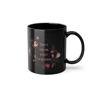 Pode incluir: Caneca de café de cerâmica preta com um design floral e o texto "Não me pergunte o que estou lendo" em rosa.