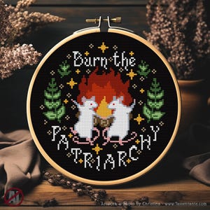 Könnte beinhalten: Ein Kreuzstich-Stickrahmen mit schwarzem Hintergrund. Das Design zeigt den Text "Burn the Patriarchy" mit zwei weißen Mäusen, die um ein Lagerfeuer tanzen. Grüne Blätter und goldene Sterne umgeben den Text und die Mäuse. Der Rahmen ist in einem Holzrahmen gefasst.