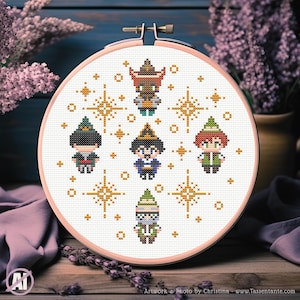 May include: A cross-stitch embroidery hoop featuring six pixelated figures in various outfits, surrounded by gold starbursts. The figures are in hats and robes, with details in red, green, blue, and brown. The hoop is set against a wooden surface.