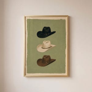 Könnte beinhalten: Ein gerahmter Kunstdruck mit einer vertikalen Anordnung von drei Cowboyhüten vor einem einfarbigen salbeigrünen Hintergrund. Der obere Hut ist schwarz, der mittlere Hut ist cremefarben und der untere Hut ist braun. Jeder Hut hat ein schmales Band.