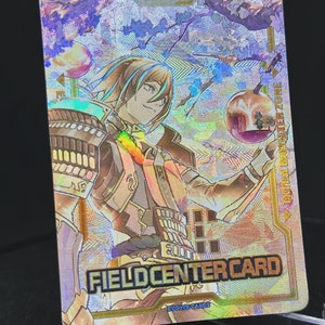 May include: A holographic trading card featuring a character with blue hair and a red and gold outfit. The card has a gold border and the text "FIELD CENTER CARD" and "EVOLVE CARDS".