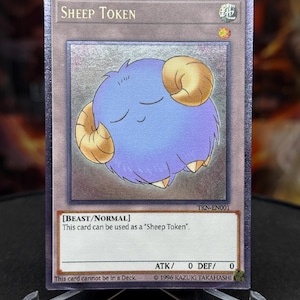 May include: A collectible card with a blue sheep character and golden horns, labelled "Sheep Token". The card has text indicating it's a Beast/Normal card. The card has a holographic sheen and is displayed on a clear stand. The card is a collectible item.