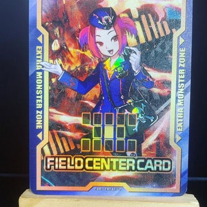 Peut inclure: Une carte de centre de terrain bleue et blanche avec une femme en uniforme bleu et un chapeau rouge tenant un appareil photo. La carte porte le texte "FIELD CENTER CARD" et "EXTRA MONSTER ZONE".