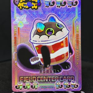 Op de afbeelding: Een holografische ruilkaart met een cartoon kattenpersonage ontworpen als een koffiekopje. De kaart heeft een paarse achtergrond met de tekst "Fun Junky Heroes" en "Field Center Card". De kattenbeker heeft rood-witte strepen.