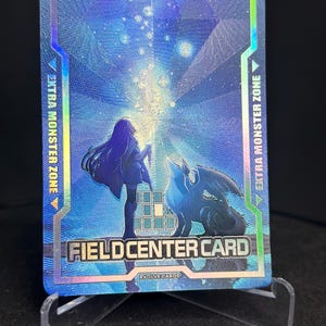 May include: A holographic trading card with a blue and purple background. The card features a silhouetted figure and a winged creature. Text on the card reads "EXTRA MONSTER ZONE" and "FIELD CENTER CARD". The card is displayed on a clear stand.