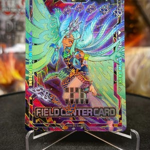 Può includere: Una carta collezionabile olografica con un'illustrazione in stile anime di una donna con capelli turchesi e un abito dettagliato. La carta mostra il testo "FIELD CENTER CARD" e "EXTRA MONSTER ZONE". Presentata su un supporto trasparente.