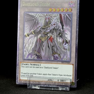 Peut inclure: Une carte Darklord Token avec une finition argentée. La carte présente une illustration détaillée d'une figure sombre avec des ailes, une armure et une épée. Le texte comprend "DARKLORD TOKEN" et les détails de la carte.