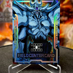 Op de afbeelding: Een holografische ruilkaart met een blauw monster met een gedetailleerd ontwerp. De kaart leest "FIELD CENTER CARD" en "EXTRA MONSTER ZONE". De kaart wordt gepresenteerd op een transparante standaard.