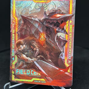May include: A trading card featuring a character from the anime series "Berserk". The card has a red, orange, and yellow background with a holographic effect. The character is Guts, who is wielding a large sword. The card also features the text "FIELD CEL" and "EVOLVE CARDS".