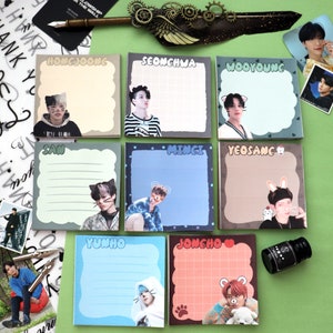 May include: Eight colourful square sticky note pads with different K-pop idols on each. The note pads have lined paper and are decorated with cartoon animals. The note pads are arranged in two rows of four. The note pads are on a green surface.