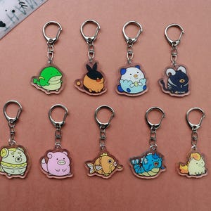 Puede incluir: Una colección de nueve llaveros de acrílico con ilustraciones coloridas de criaturas estilo dibujos animados. Cada dije está sujeto a un broche de langosta y una anilla de metal plateado. Los diseños incluyen varios animales y personajes.