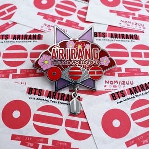 Può includere: Una spilla in smalto metallico a forma di ventaglio con sfondo rosso, accenti floreali rosa e il testo ARIRANG BTS 2026 WORLD TOUR. Sotto il ventaglio sono visualizzate tre icone circolari rosse e un piccolo ciondolo grigio pende dal centro.