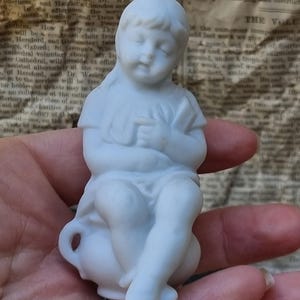 May include: A small, white porcelain figurine of a seated child with closed eyes, wearing a simple dress. The figurine is held in a hand, with a vintage newspaper in the background.