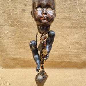 May include: A vintage-style doll head with a distressed appearance, empty eye sockets, and attached arms. It hangs from a twisted cord, with a pendant featuring coins and beads, and a heart-shaped ornament. The overall aesthetic is dark.