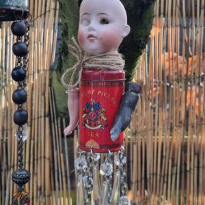 May include: A handcrafted wind chime with a vintage doll's head, a red tea tin with a crest, and crystal embellishments. The doll's head is secured to the tin with twine, and a small arm and hand are attached. The chime is suspended from a chain, creating a decorative piece.