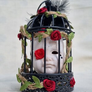 May include: A decorative black metal cage with a white face mask inside. The cage is decorated with red fabric roses, green leaves, and twine. Black feathers are attached to the top. The mask has a red rose over one eye.
