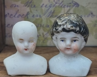 May include: Two antique porcelain doll heads. One head is bald, the other has black hair. Both have painted facial features, including blue eyes and red lips. The heads are white with some discoloration and are displayed on a wooden surface.