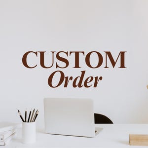 May include: A white desk featuring a silver laptop, a white pencil holder with several pencils, and a stack of white books. The wall above the desk displays the brown text CUSTOM Order in a serif font, accompanied by a gold desk lamp.