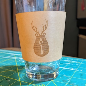 May include: A brown leather beer koozie with a laser-engraved design of a bear wearing sunglasses and a tie with antlers.