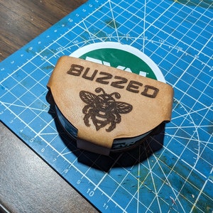 May include: A brown leather coaster with a bee design and the word "BUZZED" engraved on it. The coaster is sitting on a blue cutting mat.