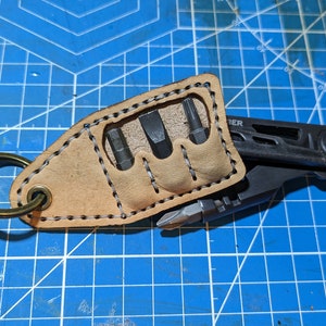 May include: A tan leather keychain with a key ring and a multi-tool with a set of screwdriver bits. The keychain is stitched with black thread and has a slot for the screwdriver bits.