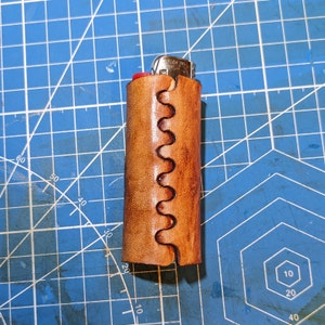Peut inclure: Un étui à briquet en cuir marron avec un motif de couture décoratif. L'étui est conçu pour contenir un briquet de taille standard.