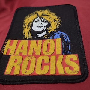 Peut inclure: Un écusson rectangulaire en tissu noir présentant une illustration graphique d'une personne aux cheveux blonds volumineux. Sous la figure, le texte HANOI ROCKS est imprimé en lettres capitales jaunes sur un fond rectangulaire rouge.