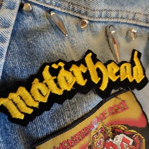 May include: Close-up of a light blue denim jacket adorned with silver cone studs and patches. The patches feature the band names "Motorhead" in yellow and "AC/DC" with a monster graphic, showcasing a rock and roll theme.