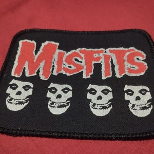 Può includere: Una toppa rettangolare in tessuto nero con bordo cucito. La parola MISFITS è stampata in un carattere rosso frastagliato nella parte superiore. Sotto il testo, quattro grafiche di teschi bianchi identiche sono disposte in fila.