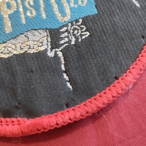 May include: Close-up of a circular patch with the text "X PISTOLS" in gold lettering on a teal and black background. The patch has a red stitched border and is on a red fabric surface.