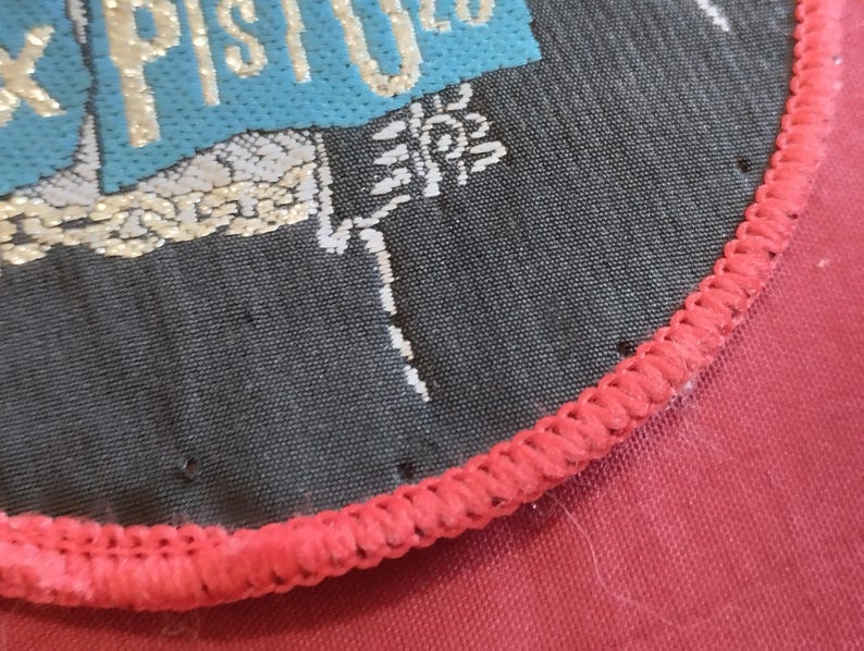 May include: Close-up of a circular patch with the text "X PISTOLS" in gold lettering on a teal and black background. The patch has a red stitched border and is on a red fabric surface.