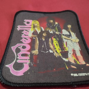 Pode incluir: Um emblema de tecido retangular com um gráfico da banda de rock Cinderella. O design inclui o nome da banda em letras cor-de-rosa grandes à esquerda e uma imagem a cores dos membros da banda. O emblema tem uma borda preta costurada.