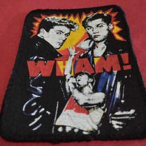 Puede incluir: Un parche de tela rectangular con un estampado gráfico del dúo musical Wham! y el texto WHAM! en letras rojas llamativas. El diseño incluye dos figuras con chaquetas negras sobre un fondo de estallido amarillo y una tercera figura debajo.