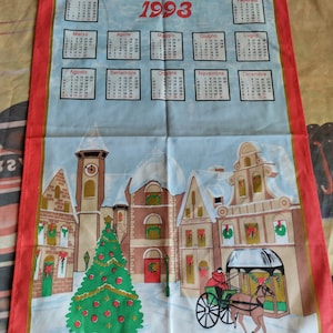 Puede incluir: Un paño de cocina vintage de 1993 con un calendario y una escena festiva de un pueblo invernal. El paño presenta un borde rojo, un fondo azul claro y una ilustración detallada de edificios, un árbol de Navidad y un carruaje tirado por caballos. Los meses del calendario están en italiano.