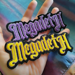 May include: Two embroidered patches with the word "Megadeth" in a gothic font. One patch features purple stitching on a black background, the other has yellow stitching. The patches are held in a hand.