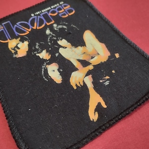 May include: A rectangular black fabric patch featuring a graphic print of the band The Doors. The design includes the text The Doors in a stylized font at the top, with orange and yellow figures printed against the black background. The edges are stitched.