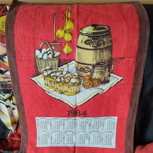 Puede incluir: Una toalla de cocina roja con borde marrón presenta un calendario de 1984 con meses y días de la semana en italiano. La toalla también tiene una imagen de bodegón de un barril, una jarra, una cesta de huevos y una cesta de champiñones.
