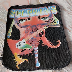 May include: A rectangular black fabric patch featuring the word SCORPIONS in white and blue gradient lettering at the top. The graphic depicts a stylised red scorpion with human-like eyes playing a white electric guitar, with a smaller yellow scorpion below.