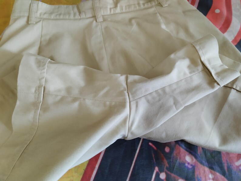 Puede incluir: Primer plano de unos pantalones cortos beige con pu&ntilde;os doblados. Los pantalones cortos tienen un dise&ntilde;o cl&aacute;sico con cierre de bot&oacute;n y cremallera. La tela parece ser un material ligero, adecuado para el clima c&aacute;lido. Los pantalones cortos est&aacute;n cuidadosamente doblados.