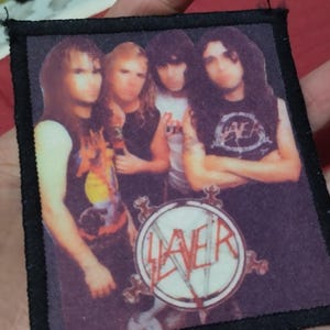 May include: A rectangular fabric patch featuring a printed image of the band Slayer with four members standing together. The patch has a black stitched border and displays the band name Slayer in red text within a circular logo at the bottom center.