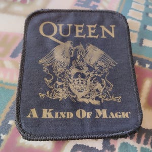 Könnte beinhalten: Ein rechteckiger schwarzer Stoffaufnäher mit dem goldfarbenen Queen-Bandlogo, dem Schriftzug "QUEEN" oben und "A KIND OF MAGIC" unten. Das zentrale Design zeigt ein detailliertes Wappen mit Flügeln, einer Krone und kunstvollen Verzierungen.