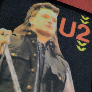 May include: A black and white patch with a photo of a man with long hair and a black leather jacket. The text "U2" is in orange and yellow on the right side of the patch.